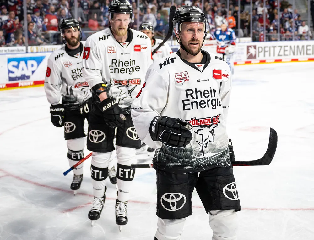 Three ice hockey players in white jerseys with black logos and graphic designs stand on the ice.