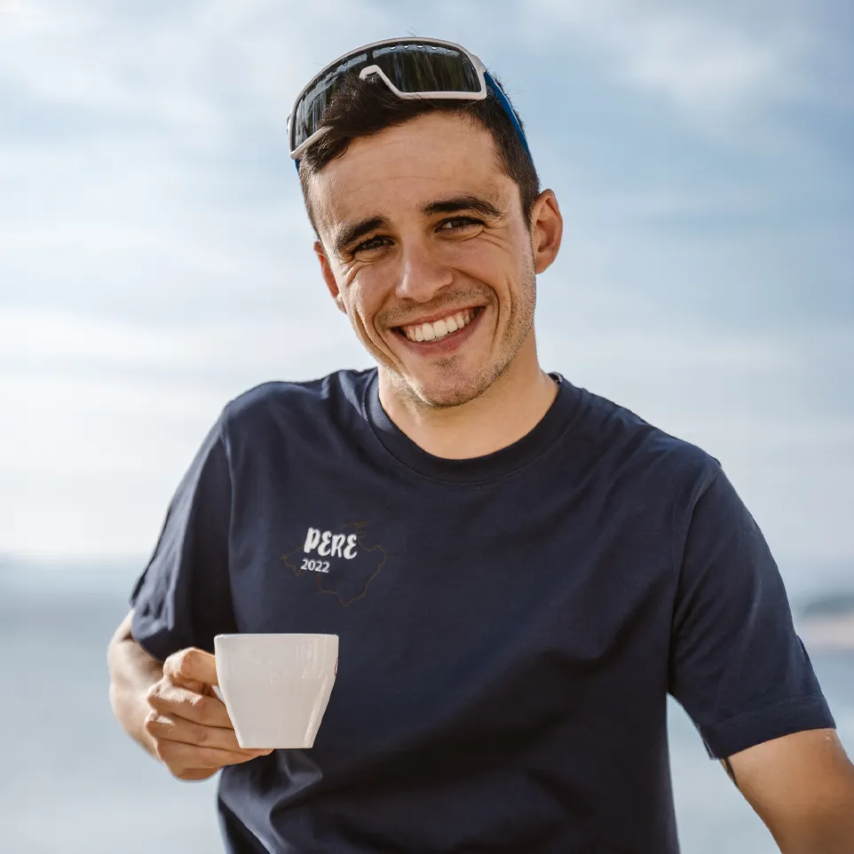 A man is holding a cup and wearing a navy blue t-shirt with the print 'PERE 2022', smiling in front of a blurred background.