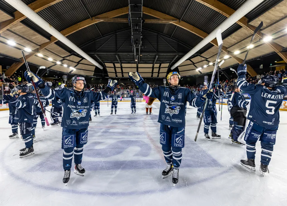 Ice hockey players in blue jerseys stand on the ice, celebrating with raised sticks in front of an cheering crowd.