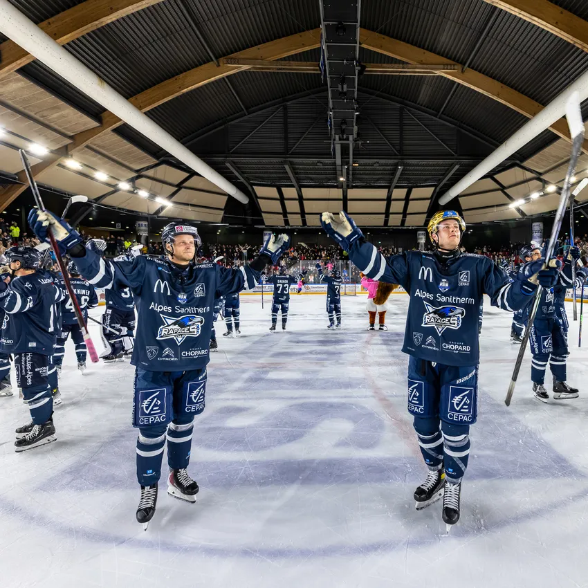 Two ice hockey players on the ice celebrating with their sticks while other players and spectators are visible in the background.