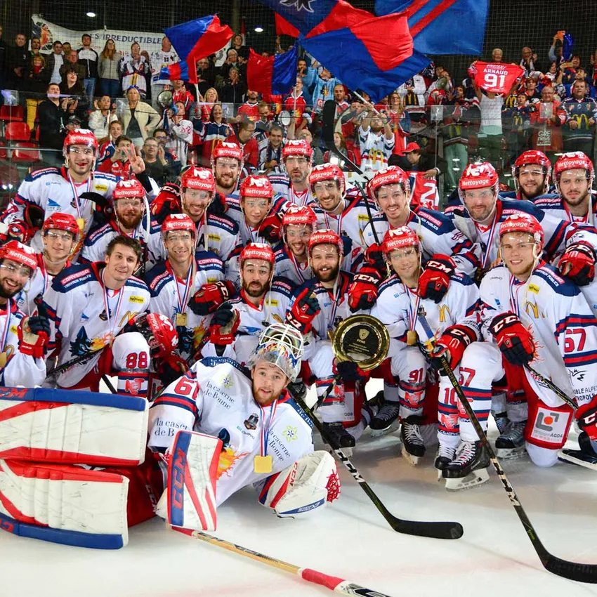 A group of ice hockey players in white jerseys with red and blue accents proudly poses with medals and a trophy on the ice.