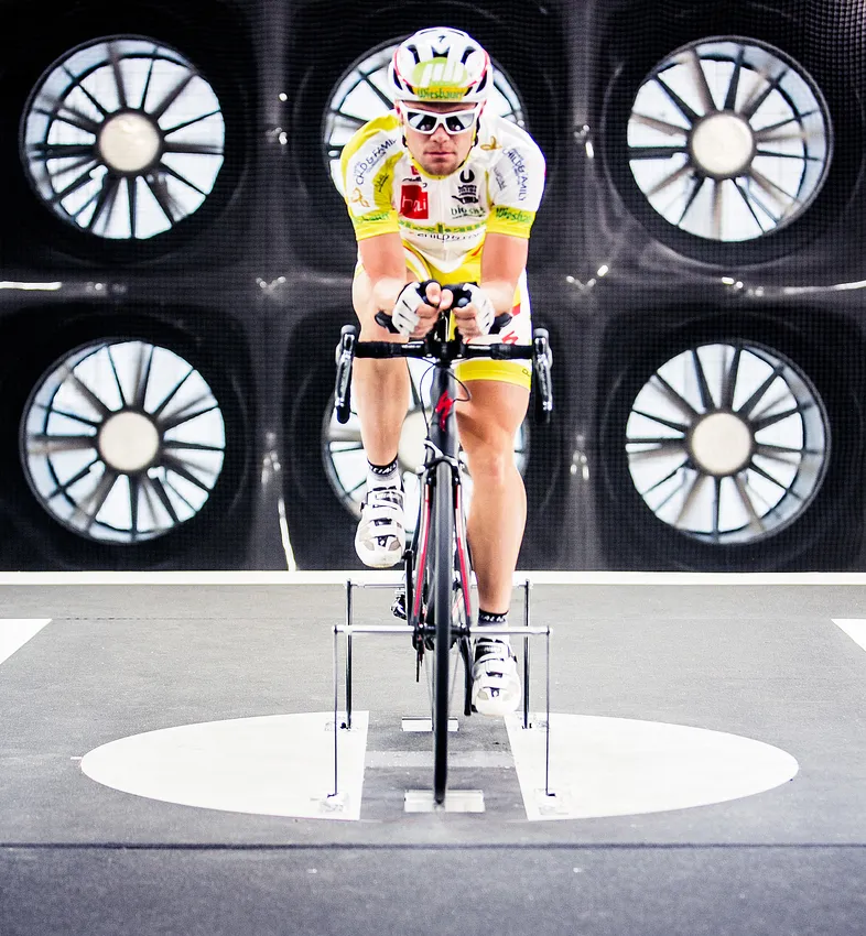 A cyclist in an aerodynamic position on a stationary bike in a wind tunnel, testing the performance of customized sportswear.