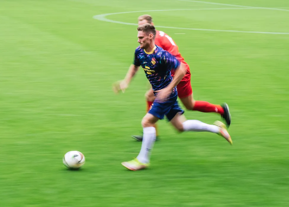 A soccer player in a blue jersey with colorful patterns runs swiftly across the green field while another player closely follows him.