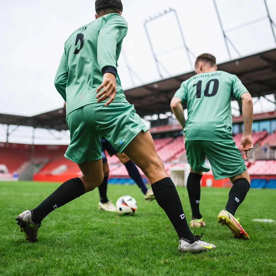 Two football players in green jerseys and black socks actively playing on a grass field in a stadium.