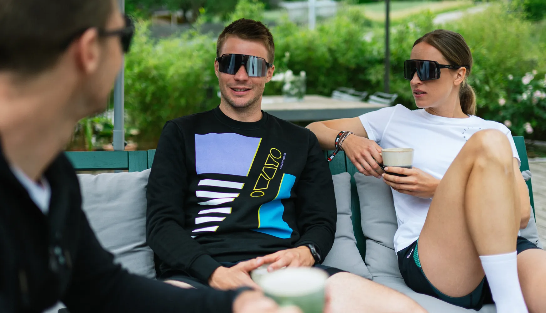 Three individuals are relaxed on a couch, one wearing a black sweatshirt with a colorful design, and the other in a white t-shirt, both holding drinks.