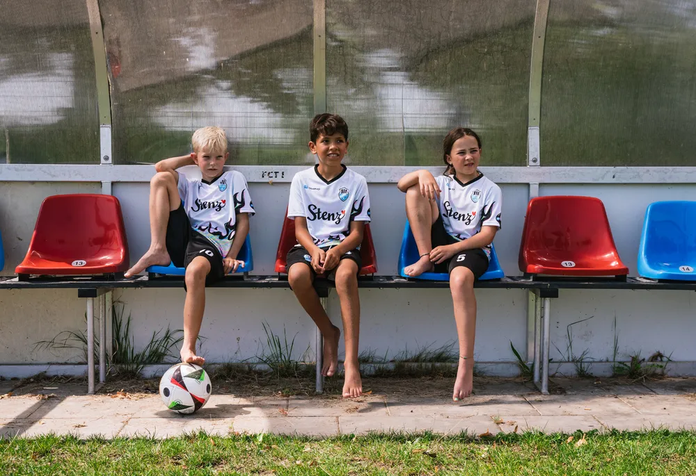 Three children in sporty soccer jerseys sit on a bench next to a soccer ball.