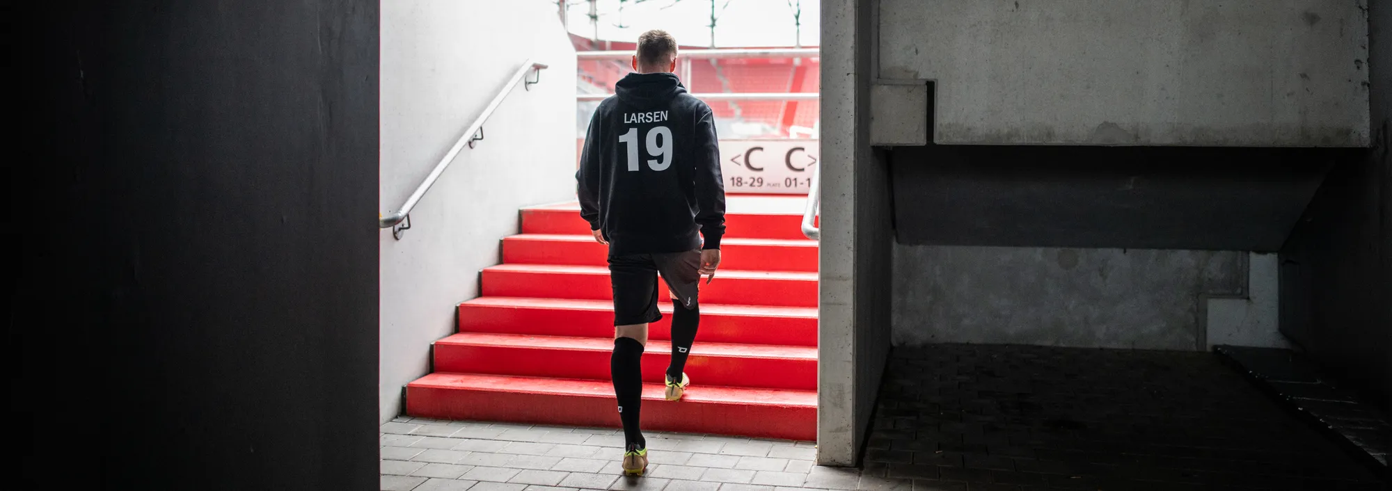A player in a dark hoodie and shorts is walking up red stairs out of a stadium.