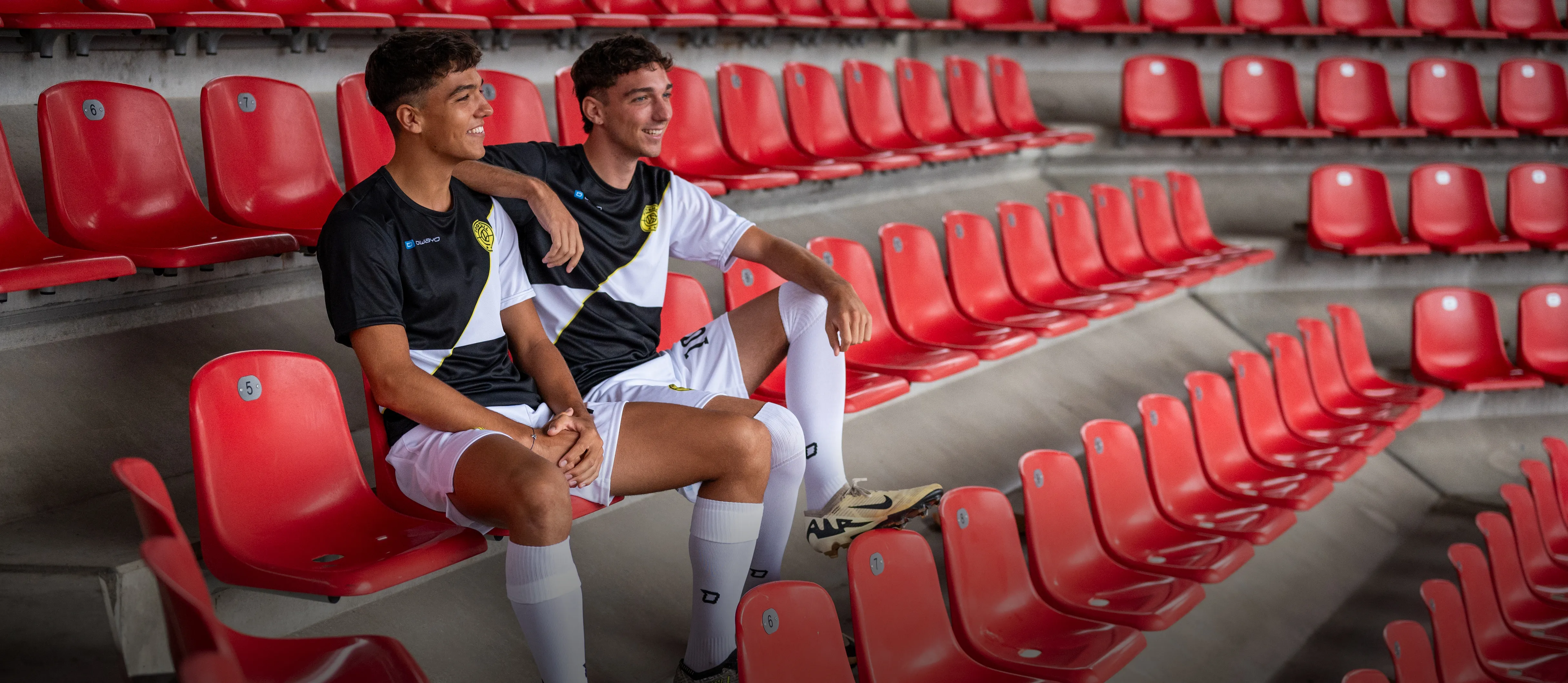 Two young men in sporty football attire are casually sitting on red stadium seats.