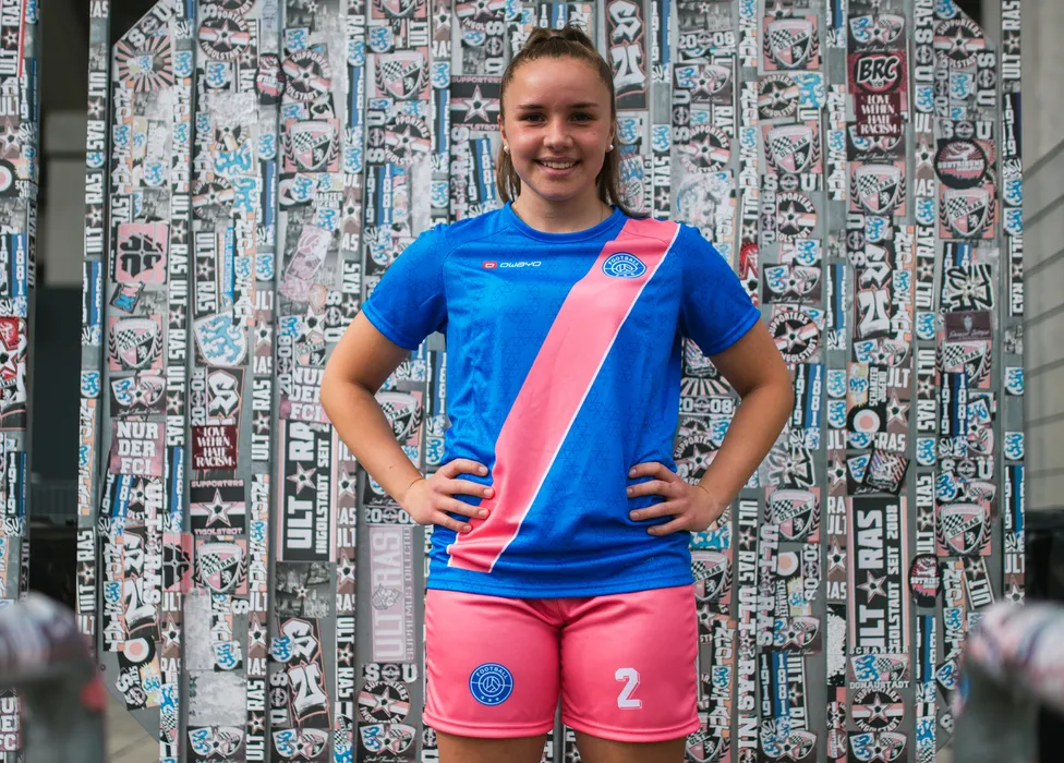 A young athlete poses in a blue soccer jersey with a pink stripe and pink shorts in front of a colorful wall covered in stickers.