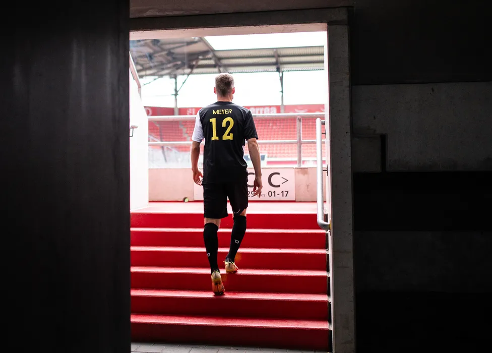 A football player wearing a black jersey with the number 12 ascends the red stairs in the stadium.