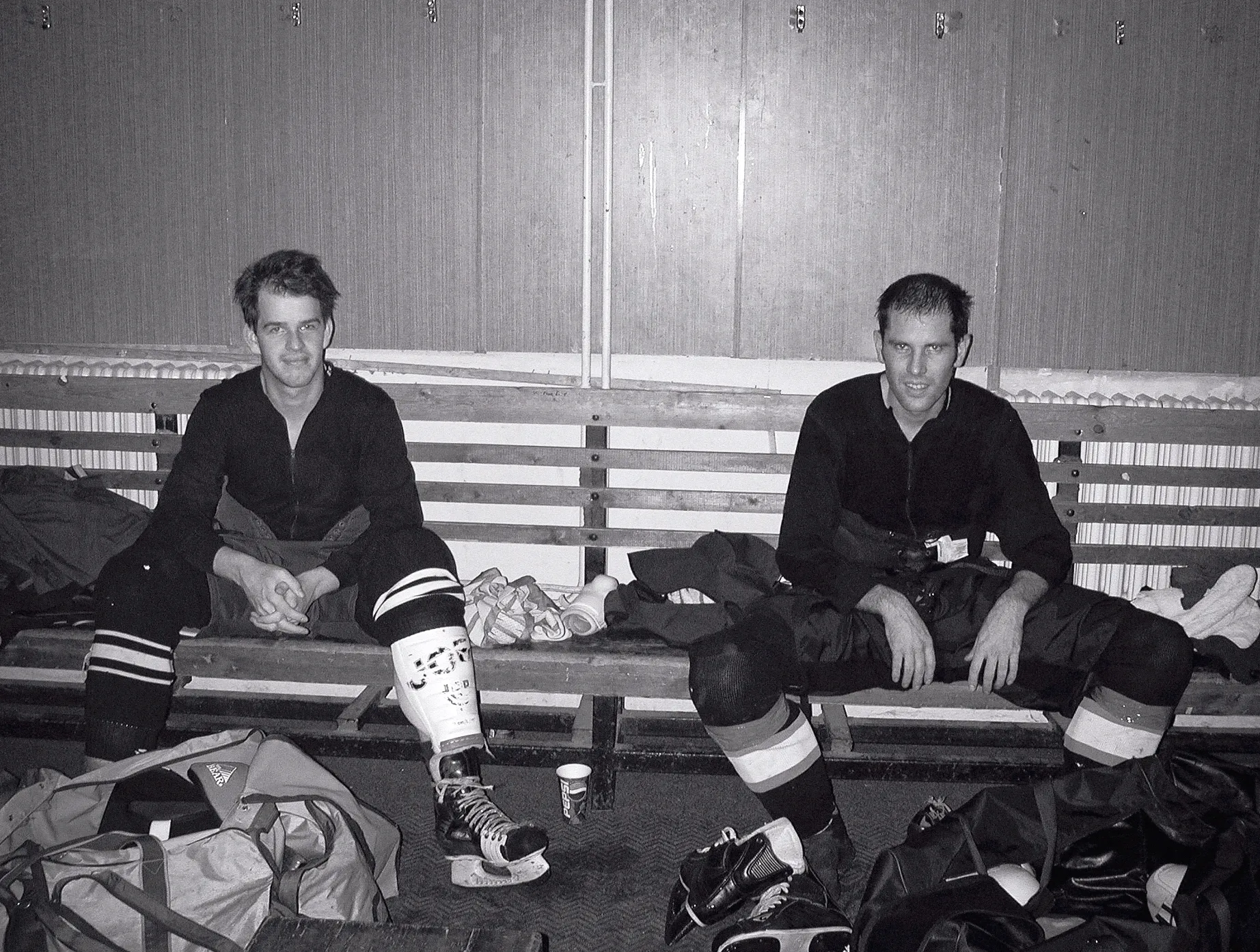 Two ice hockey players in full gear sit on a wooden bench in a locker room, surrounded by sports bags and skates.