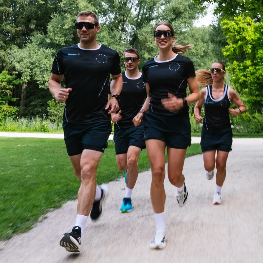 Four individuals in athletic running gear are jogging along a path through a green park landscape.