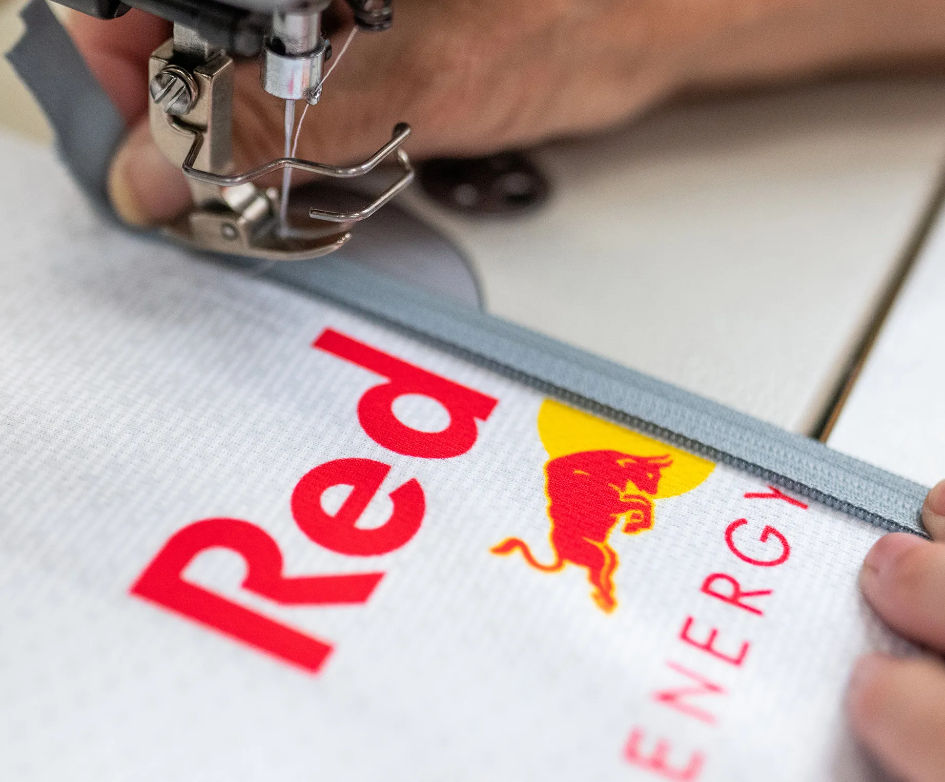A sewing machine is attaching a grey zipper to a white sportswear garment with the Red Bull logo.