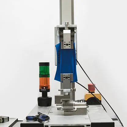 A blue fabric is being tested in a machine on a workbench, next to a signal light and tools.