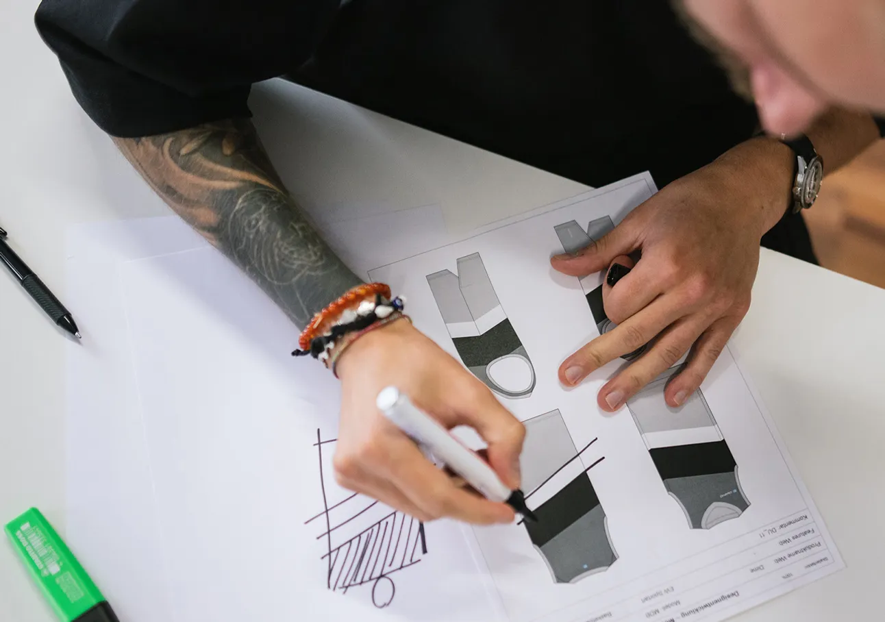 A person with a tattoo on their arm is drawing with a white marker on a sheet of paper showing several designs for sportswear.
