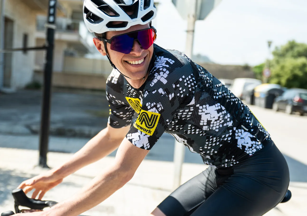 A cyclist wearing a black and white patterned cycling jersey with a yellow sponsor logo, riding on a street.