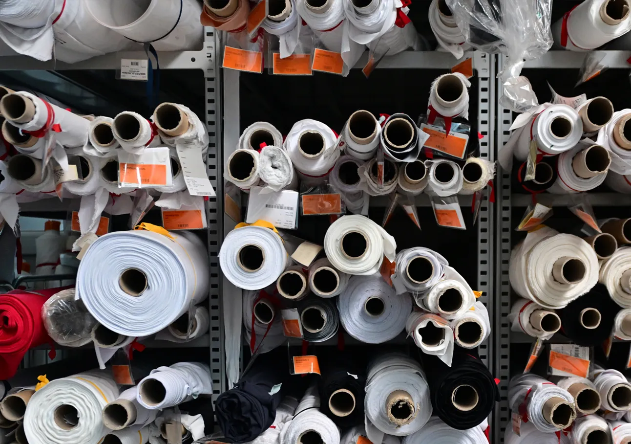 Shelves filled with rolls of fabric in various colors, including white, black, and red, with tags indicating an in-house production facility.
